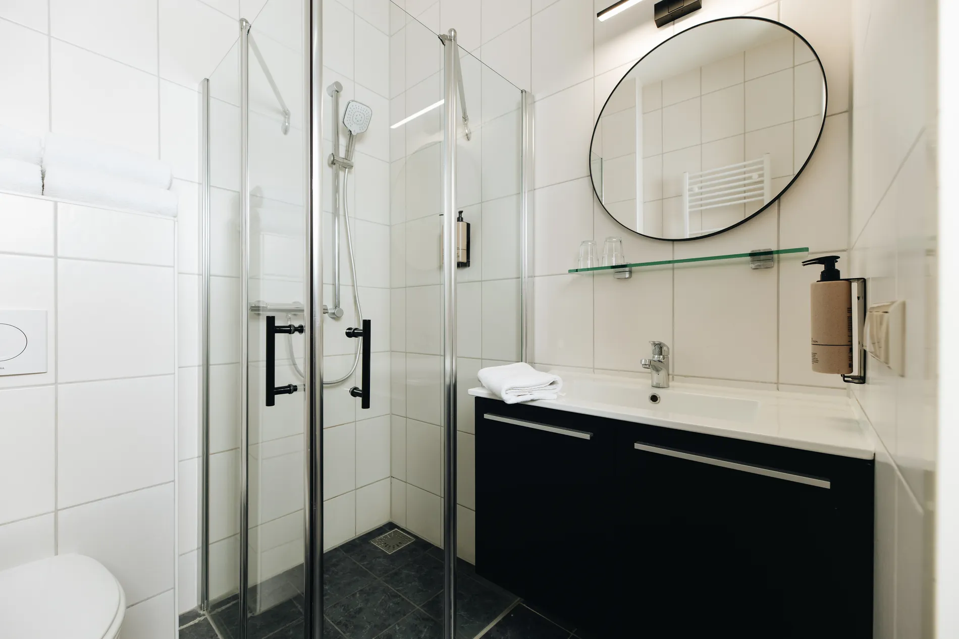 Moderne, lichte badkamer met een glazen douchecabine, een zwart wastafelmeubel met witte wasbak en een ronde designspiegel.